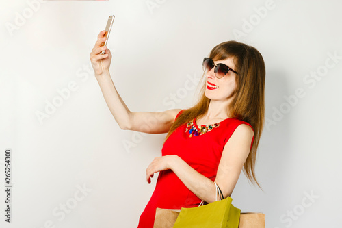 Young girl in a red dress takes pictures of herself on the phone and holds packages from stores on a light background. Online shopping concept  shopping app  sale  selfie.