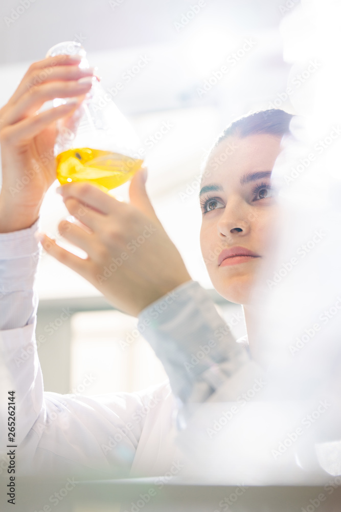 Serious pensive young female scientist standing in laboratory and holding flask with yellow liquid while researching liquid substance Serious pensive young female scientist standing in laboratory and holding flask with yellow liquid while researching liquid substance