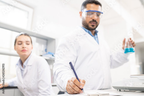 Thoughtful young multi-ethnic laboratory coworkers working on drugs in pharmaceutical company: Arabian scientist checking liquid substance and making records in clipboard