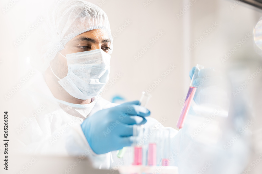 Serious concentrated young Arabian medical scientist in protective workwear and mask comparing liquids in test tubes while working with hazardous substances in laboratory Serious concentrated young Arabian medical scientist in protective workwear and mask comparing liquids in test tubes while working with hazardous substances in laboratory