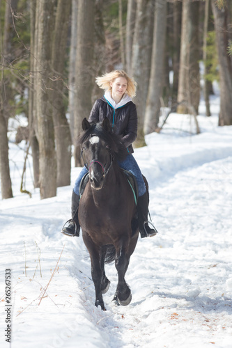 A winter forest. A young blonde woman riding a horse on snowy ground