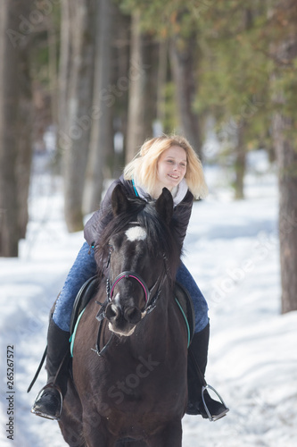 A winter forest. A young smiling woman riding a black horse on snowy ground