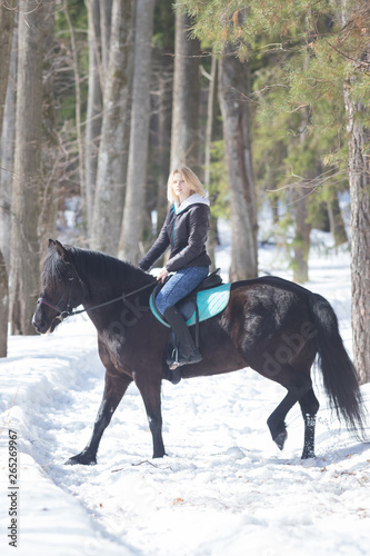 A young blonde woman riding a black horse in snowy forest