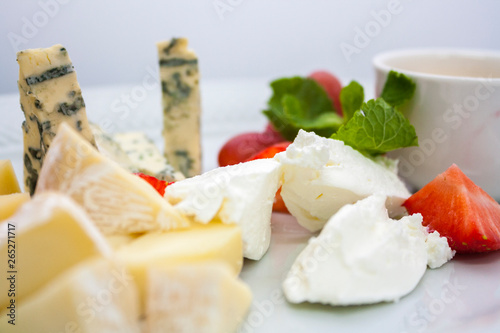 a plate of several kinds of cheeses in assortment, with strawberries and grapes, on a white plate on a white background, close-up