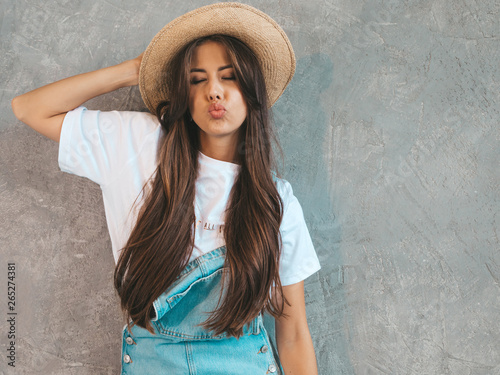 Young beautiful woman looking at camera. Trendy girl in casual summer overalls clothes and hat. Funny and positive female posing near gray wall in studio. Making duck face