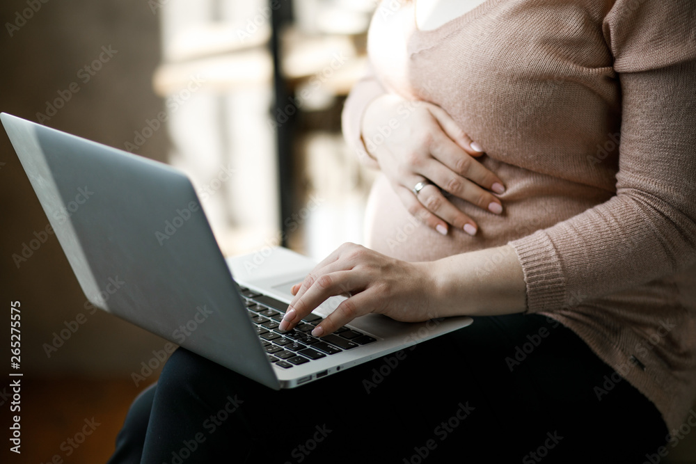 Naklejka premium Pregnant woman working on laptop. Cropped image of pregnant businesswoman typing something on laptop while sitting at her working place in office