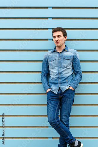 Growth portrait of brunet cute young man in casual stylish denim wear. Put his hands in his pockets. He leaned against blue wooden wall.