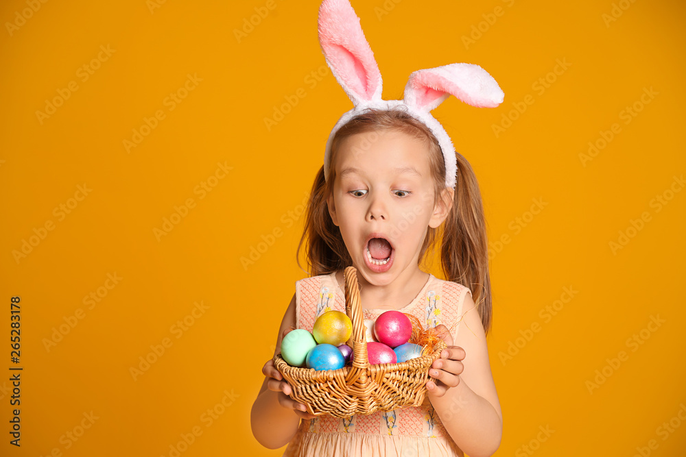Surprised little girl with basket of Easter eggs and bunny ears on color background - obrazy, fototapety, plakaty 