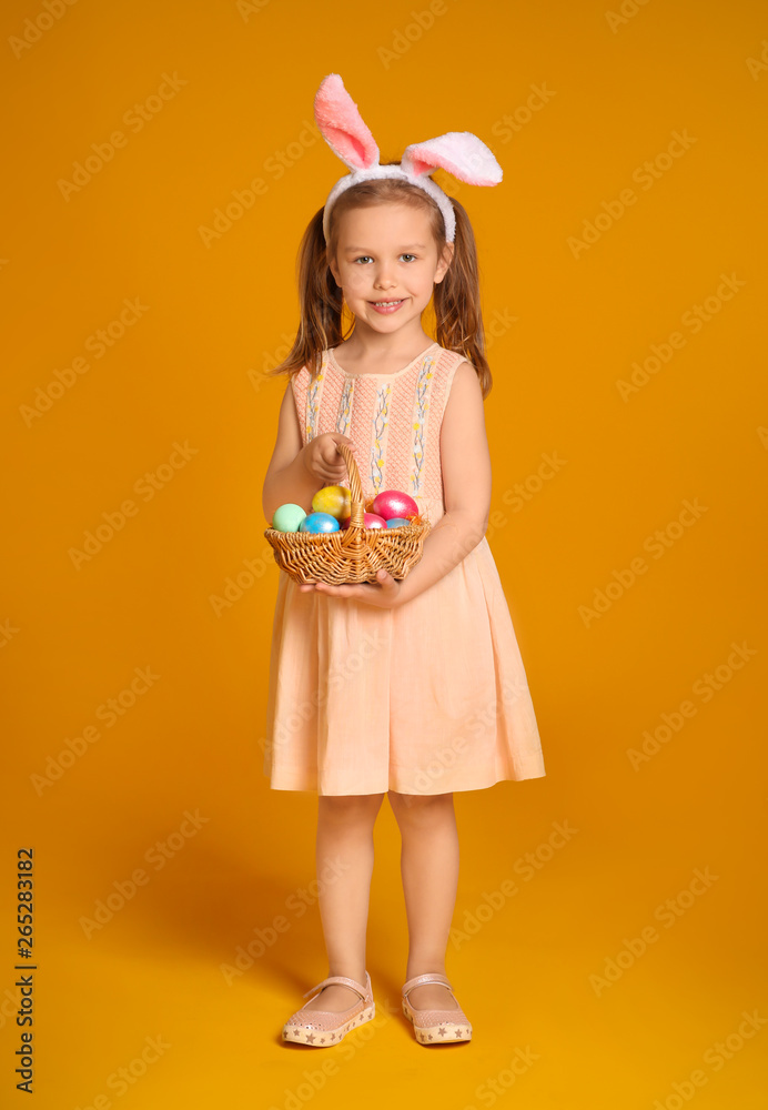 Little girl with basket of Easter eggs and bunny ears on color background
