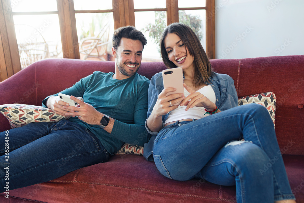 Lovely young couple using they mobile phone while sitting on sofa at home.