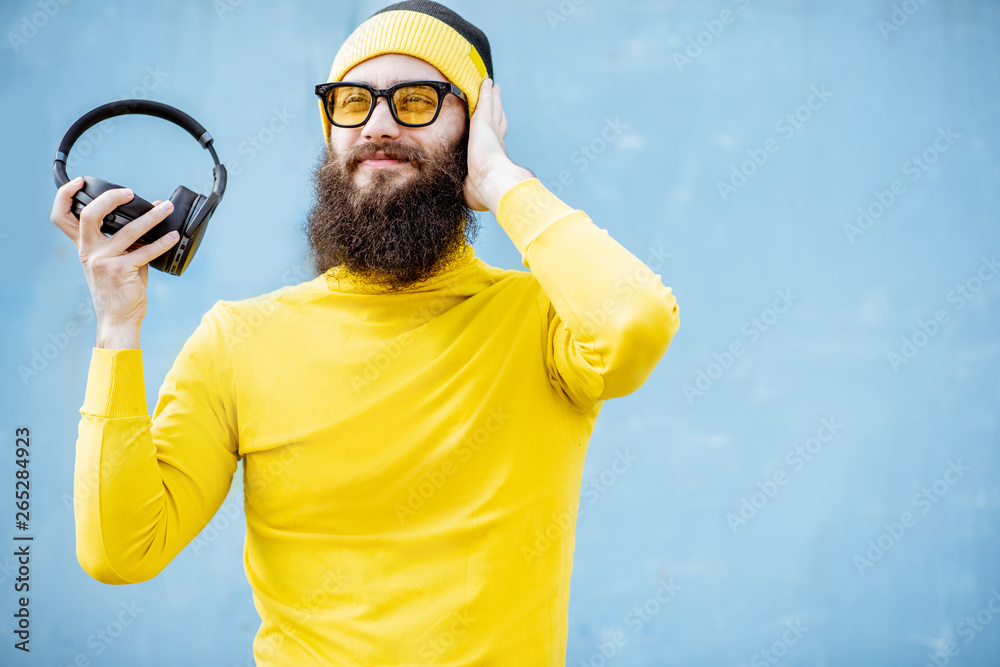 Portrait of a stylish bearded man in yellow clothes enjoying music with headphones on the blue background Portrait of a stylish bearded man in yellow clothes enjoying music with headphones on the blue background