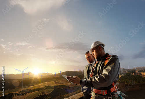 Windmill engineer and businessman planing new ecology project. they standing and looking in tablet. Around wind generators blueprint and beautiful landscape