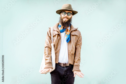 Portrait of a stylish man dressed in jacket, hat and scarf standing with bag on the turquoise background outdoors