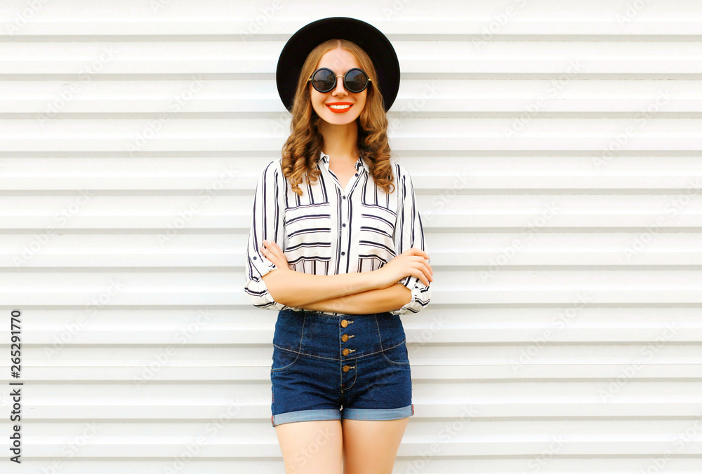 Happy smiling young woman in black round hat, shorts, white striped shirt posing on white wall background