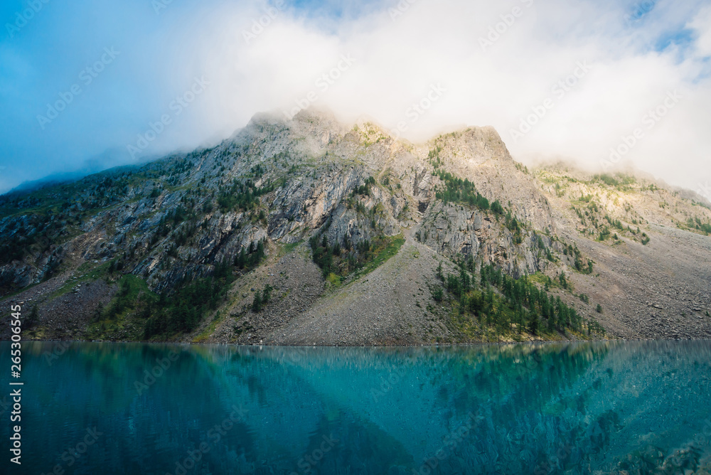 Giant cloud above rocky ridge with trees in sunlight. Amazing mountain lake. Mountain range under blue cloudy dawn sky. Wonderful rocks. Morning landscape of highland nature. Low clouds in sunny light