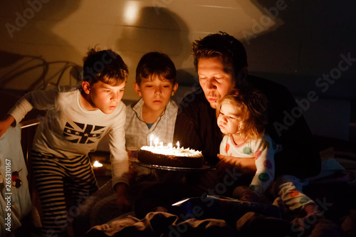 Three children, toddler girl and two school kids boys congratulating father to his birthday. Baby sister child, two brothers and dad with cake blowing candles. Happy healthy family portrait