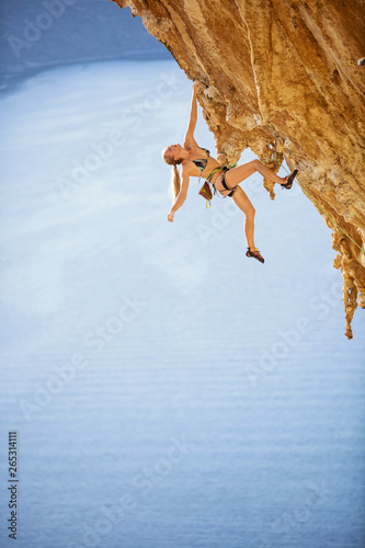 Young female rock climber in bikini hanging with one hand on overhanging cliff
