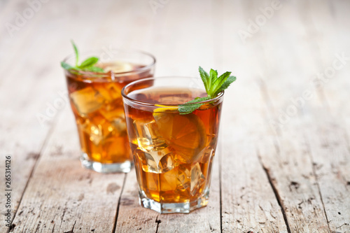 Two glasses with traditional iced tea with lemon, mint leaves and ice cubes in glass on rustic wooden table background.
