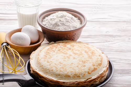Stack of French crepes in frying pan on wooden kitchen table with milk eggs and flour aside