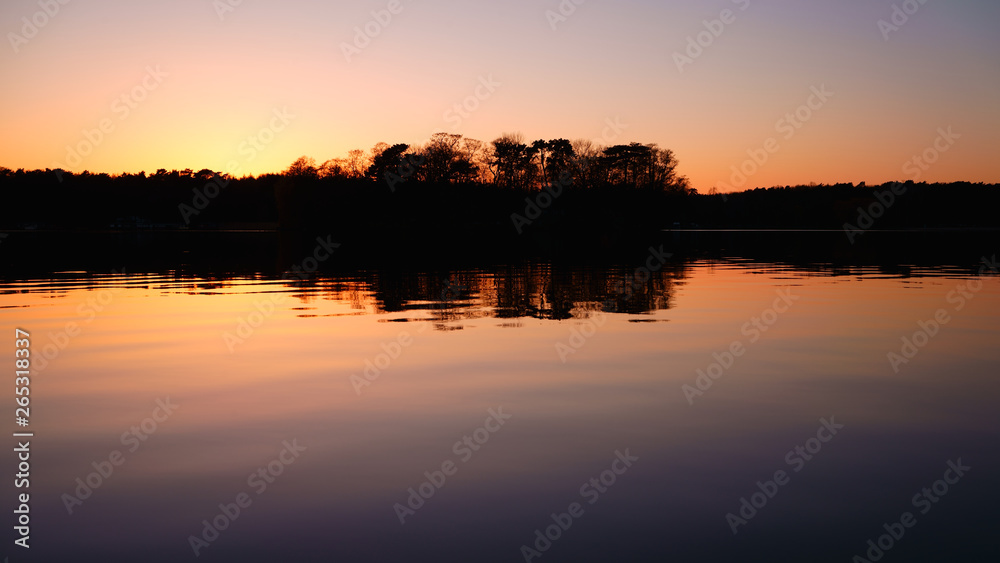 Romantic sunset over the lake. The silhouette of the trees reflects in the calm waters. Colorful landscape background, atmospheric and tranquil. 