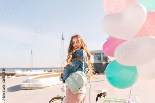 Smiling brunette girl came to the pier to let the colorful balloons into the blue sky. Portrait of gorgeous young long-haired woman with white bicycle posing in front of the sea after birthday party.