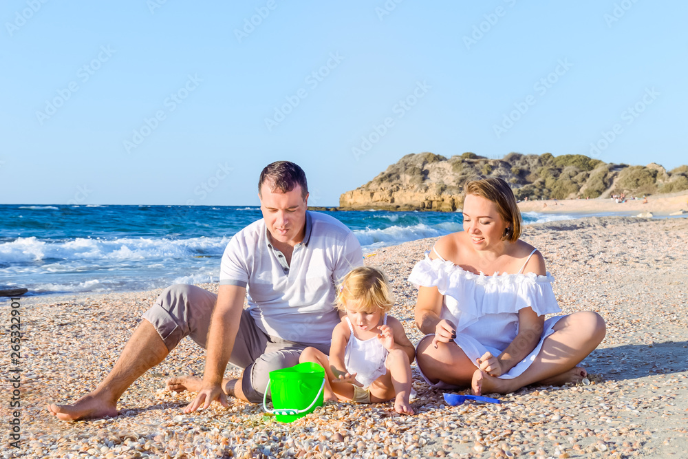 Happy family of three - pregnant mother, father and daughter having fun, playing with sand and shells on the beach. Family vacation, travel concept. Selective focus. Copy space.