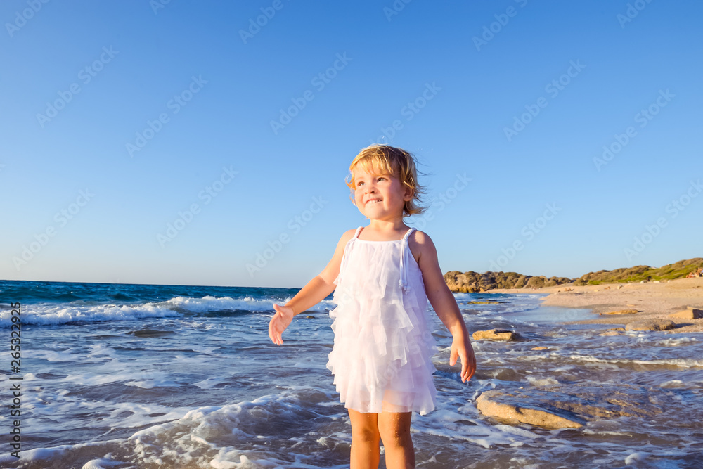 Portrait of cute little adorable toddler girl in white clothes walking on the empty beach on a warm sunny summer day. Holidays at sea. Family vacation. Selective focus, space for text. Portrait of cute little adorable toddler girl in white clothes walking on the empty beach on a warm sunny summer day. Holidays at sea. Family vacation. Selective focus, space for text.