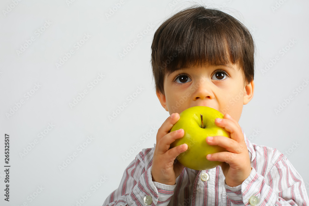 Small caucasian boy with dark hair, big dark eyes, wearing striped shirt is eating, biting big green apple, isolated on white background Small caucasian boy with dark hair, big dark eyes, wearing striped shirt is eating, biting big green apple, isolated on white background