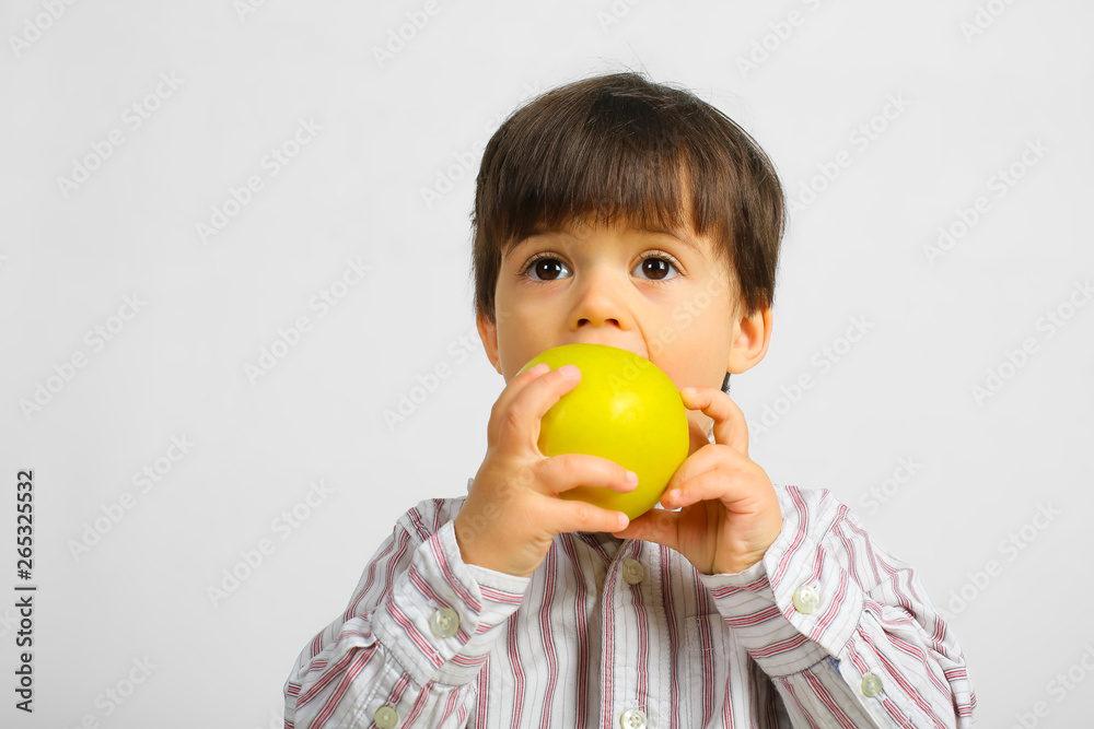 Small caucasian boy with dark hair, big dark eyes, wearing striped shirt is eating, biting big green apple, isolated on white background Small caucasian boy with dark hair, big dark eyes, wearing striped shirt is eating, biting big green apple, isolated on white background