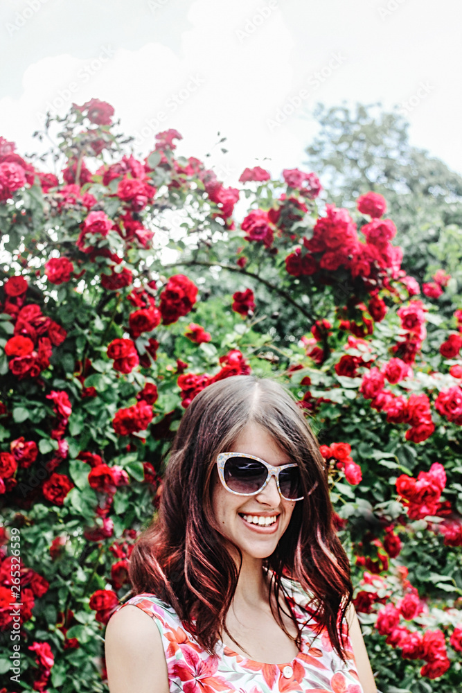 stylish brunette beautiful woman smiling in floral dress and retro sunglasses on background of sunny roses stylish brunette beautiful woman smiling in floral dress and retro sunglasses on background of sunny roses