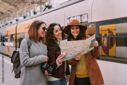 Three women travelers friends are at the train station checking the map of the city. They are laughing together while looking for a place to go. Travel concept