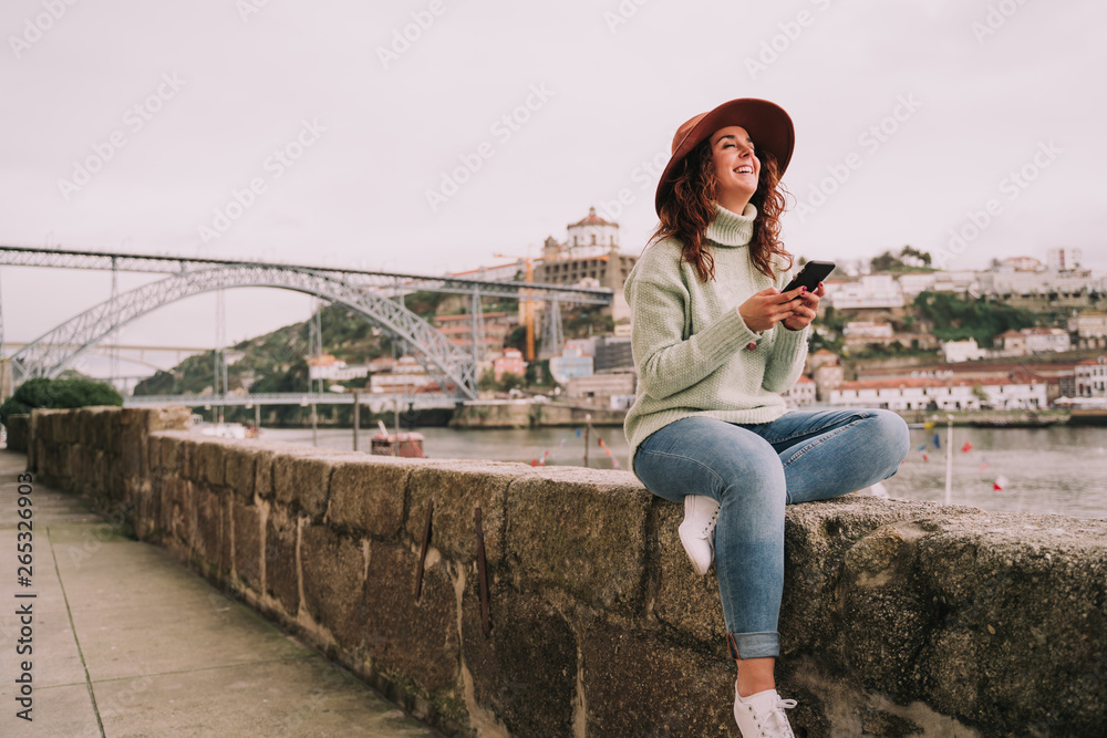 A beautiful young woman traveler is enjoying sightseeing in Porto. She is sitting on a wall near the bridge while laughing using her mobile phone. Lifestyle