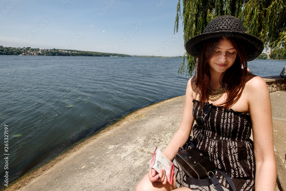 happy stylish woman sitting  at sunny shore near water, summer travel concept, space for text