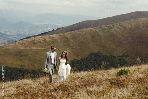 happy gorgeous bride and groom walking  in sun light holding hands  boho wedding couple  luxury ceremony at mountains with amazing view  space for text
