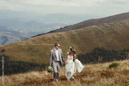 happy gorgeous bride and groom walking  in sun light holding hands, boho wedding couple, luxury ceremony at mountains with amazing view, space for text © sonyachny