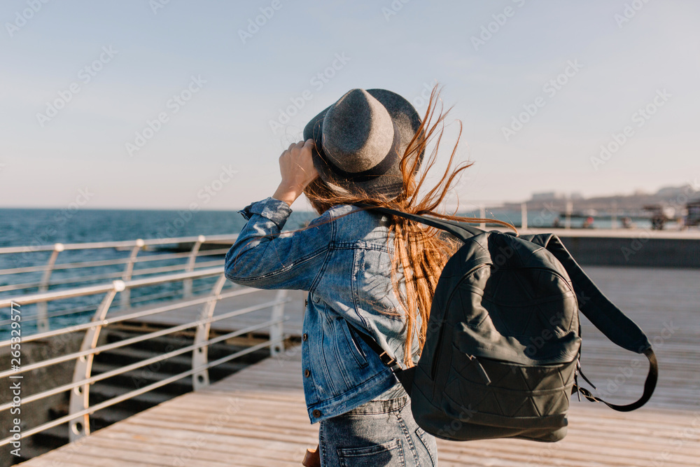 Long-haired girl in denim outfit looks into the sea far, while wind plays with her dark hair. Stylish young woman in hat with trendy backpack coming to lake to enjoy fresh air and see beautiful view