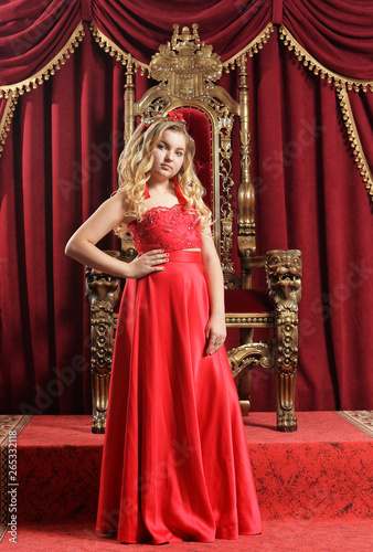 Blonde teenage girl in bright red dress standing in front of vintage chair
