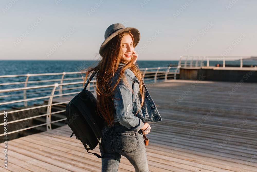 Slender long-haired girl in a denim suit strolls along the sea wharf, looking over her shoulder. Adorable brunette young woman with backpack and stylish hat enjoys morning at the ocean and smiling