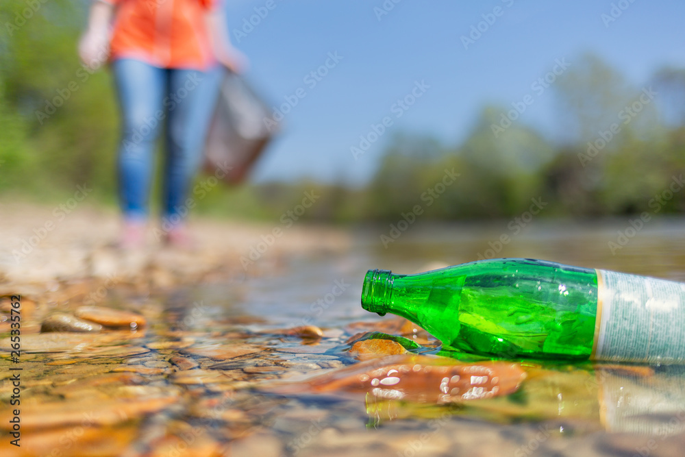 On the banks of the river is a glass bottle thrown by someone. In the background is a volunteer. The concept of environmental protection and sustainability - obrazy, fototapety, plakaty 