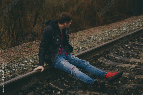Trendy young man sitting on rails Pensive male in jeans and leather jacket sitting on railway in evening of cloudy day