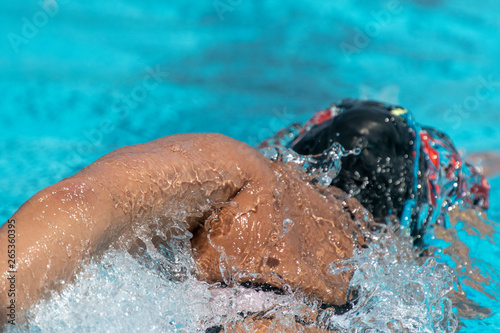 Close-up of woman swimming freestyle. Sun tanned skin. Black swim cap, white tech suit. Blue pool water on sunny day. Splashing water.