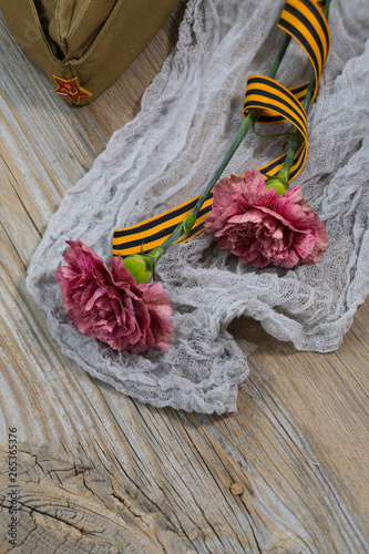 Two pink carnations, Saint George ribbon and military cap on a wooden surface.