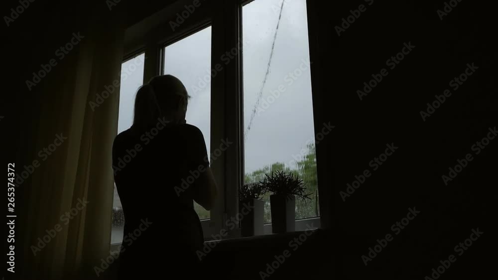 Woman standing at the window in the room crying, covering his face with his hands. It's raining outside, drops on the glass.