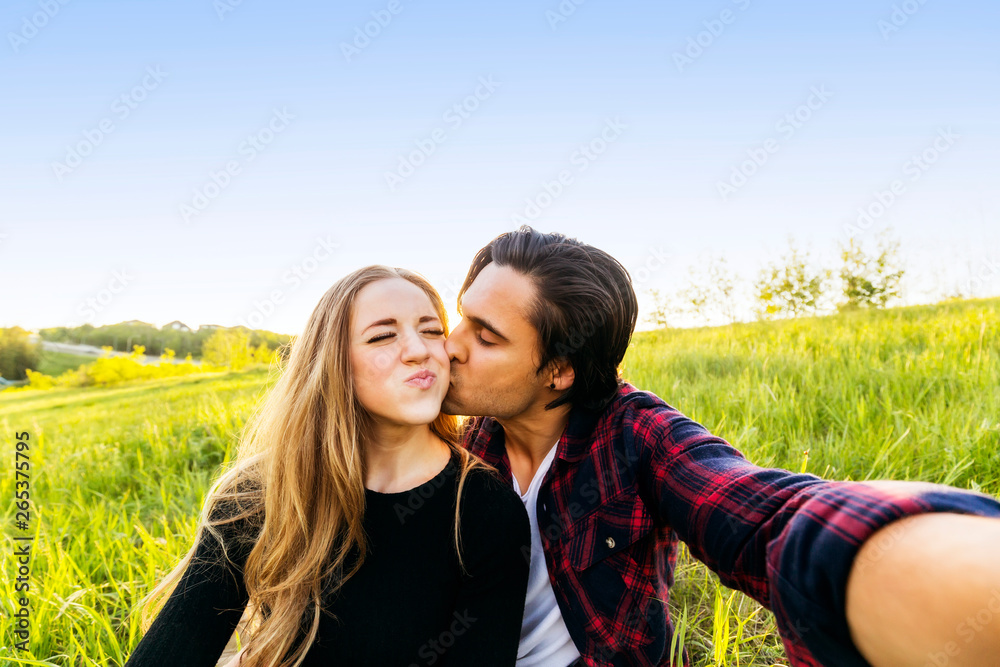 A young couple sits in a field taking a self-portrait while the young man kisses the young woman on the cheek; Edmonton, Alberta, Canada A young couple sits in a field taking a self-portrait while the young man kisses the young woman on the cheek; Edmonton, Alberta, Canada