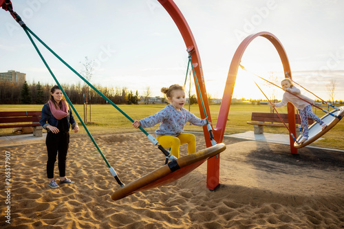 A young mom and her daughters playing on saucer swings in a playground on a warm autumn evening; Edmonton, Alberta, Canada