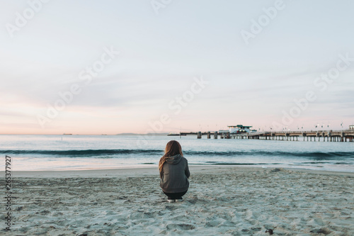A girl crouches on the beach looking out over the ocean to the horizon at sunset; Long Beach, California, United States of America