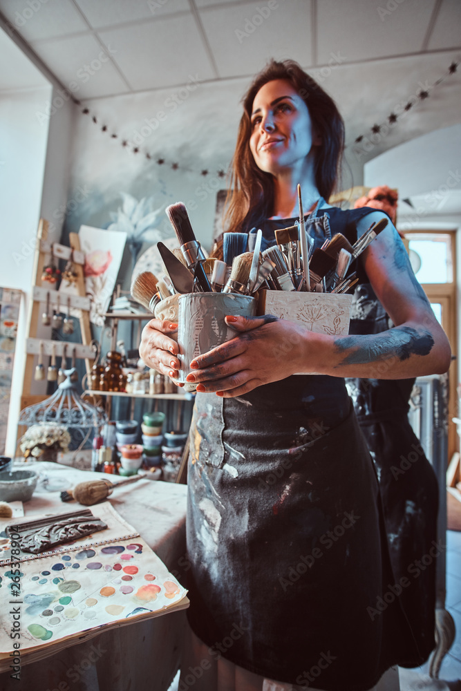 Young attractive painter is posing with brushes in her own cozy workshop.