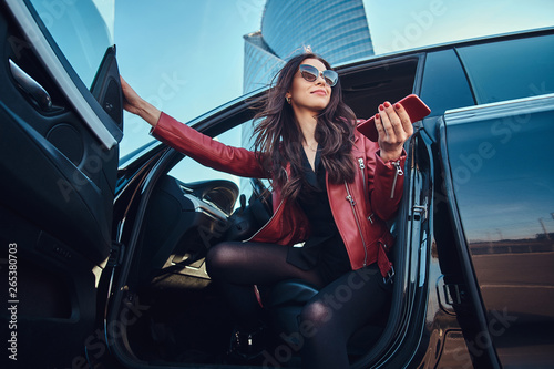 Beautiful smart women is posing in her new car while chatting on mobile phone. She is wearing red leather jacket and sunglasses.