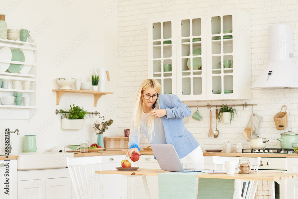 Freelance concept. Beautiful business woman in casual clothes and glasses is examining documents and smiling while working with a laptop in kitchen. Working at home Freelance concept. Beautiful business woman in casual clothes and glasses is examining documents and smiling while working with a laptop in kitchen. Working at home