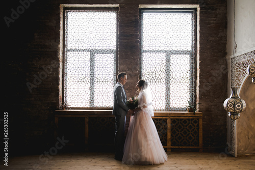 The first meeting of the bride and groom. Pink wedding dress, gray suit and stylish bouquet. Room in the style of Morocco.